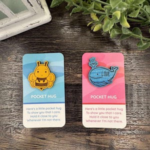 K&ouml;nnte beinhalten: Zwei Pocket-Hug-Karten. Eine Karte ist blau mit einer Holzbiene, die andere rosa mit einem Holzwal und dem Wort "LOVE". Jede Karte hat den Text "POCKET HUG" und eine Nachricht.