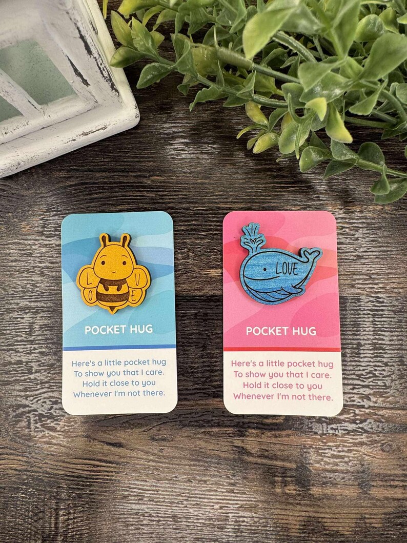 K&ouml;nnte beinhalten: Zwei Pocket-Hug-Karten. Eine Karte ist blau mit einer Holzbiene, die andere rosa mit einem Holzwal und dem Wort "LOVE". Jede Karte hat den Text "POCKET HUG" und eine Nachricht.