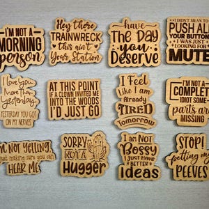 Puede incluir: Recortes de madera con frases humor&iacute;sticas grabadas, como "No soy una persona de la ma&ntilde;ana" y "Lo siento, no soy de abrazos". El texto marr&oacute;n oscuro contrasta con la madera marr&oacute;n claro. Ideal para regalos.