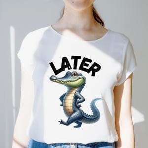 May include: White t-shirt featuring a cartoon alligator with a blue and tan body, smiling and standing with its arms crossed. The word "LATER" is printed above the alligator in bold black letters.
