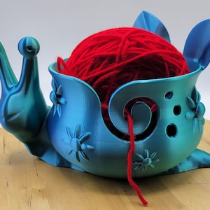 May include: A teal blue 3D printed snail yarn bowl with a red ball of yarn inside. The bowl has a hole in the side for the yarn to feed through. The snail has a detailed shell with flower designs.