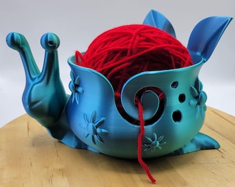 May include: A teal blue 3D printed snail yarn bowl with a red ball of yarn inside. The bowl has a hole in the side for the yarn to feed through. The snail has a detailed shell with flower designs.