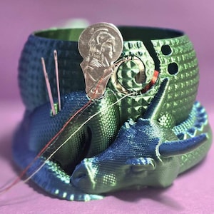 May include: A green and blue 3D printed yarn bowl with a dragon design. The bowl has a metal coin-shaped piece with a design on it. There are three needles sticking out of the bowl.