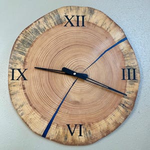 May include: A wooden clock with Roman numeral markings and blue resin accents. The clock face is a cross-section of a tree trunk, showing the growth rings.
