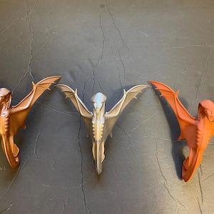 May include: Three decorative wall sculptures in the shape of a winged humanoid figure. The sculptures are made of a smooth, metallic material and are painted in different colors: copper, silver, and orange.