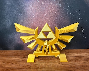 May include: A gold-colored, 3D-printed emblem featuring a stylized triangle with a smaller triangle inside, flanked by wings and crescent shapes. The emblem is set on a wooden surface with a dark, speckled background.