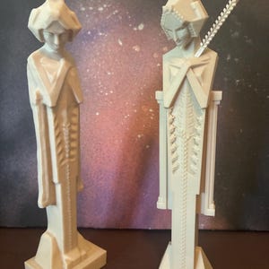 May include: Two off-white, tall, slender figurines with geometric, angular features. Each figure stands on a rectangular base, wearing a long, textured garment with a central vertical pattern. One figure has a thin, pointed protrusion extending from its back.