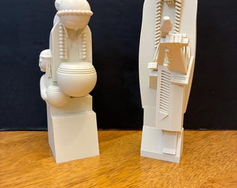 May include: Two off-white matte sculptures standing upright. The left sculpture depicts a figure with a rounded headpiece holding a bowl. The right sculpture features a tall figure with a feathered headdress and geometric details on the torso.