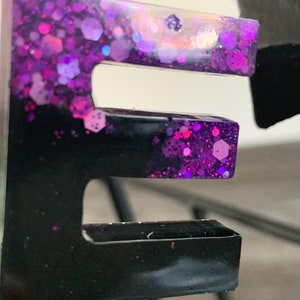 Op de afbeelding: Een zwarte en paarse harsletter E met glitter ingebed in de hars. De letter is een hanger en heeft een zilveren lus aan de bovenkant.