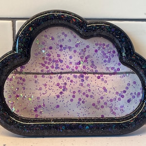 Puede incluir: Bandeja de resina con forma de nube con borde negro brillante. El centro transparente está lleno de purpurina morada. Esta bandeja decorativa se puede utilizar para guardar objetos pequeños o como decoración.