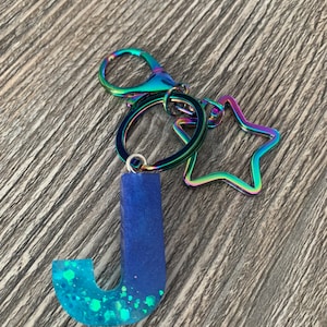 May include: A blue and green glitter resin letter J keychain with a rainbow iridescent star charm and a silver split ring.