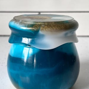 以下が含まれることがあります： 蓋付きの小さな丸い瓶。瓶は青のグラデーションで、蓋と縁に白と金のアクセントがあります。瓶は光沢のある仕上がりで、樹脂または同様の素材で作られているようです。瓶は白い表面に置かれています。