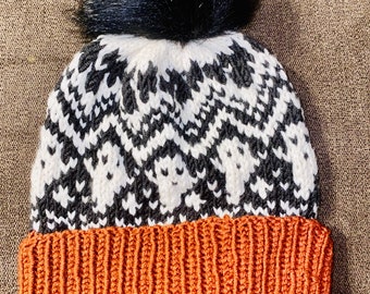 Op de afbeelding: Een witte en zwarte gebreide beanie met een zwarte pompon bovenop. De beanie heeft een bruin gebreide boord.