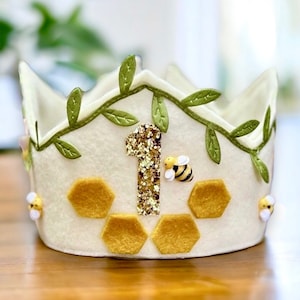 Puede incluir: Una corona de fieltro blanco decorada con hojas de fieltro verde, un n&uacute;mero uno de purpurina dorada y formas de panal de abeja de fieltro amarillo. Tambi&eacute;n hay peque&ntilde;as abejas de fieltro amarillo y negro en la corona.