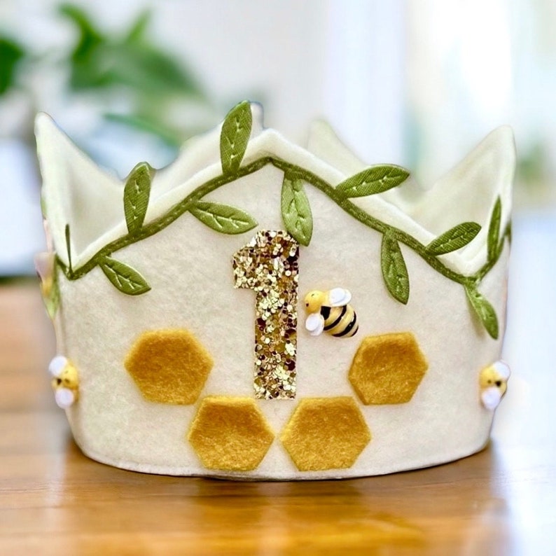 Puede incluir: Una corona de fieltro blanco decorada con hojas de fieltro verde, un n&uacute;mero uno de purpurina dorada y formas de panal de abeja de fieltro amarillo. Tambi&eacute;n hay peque&ntilde;as abejas de fieltro amarillo y negro en la corona.