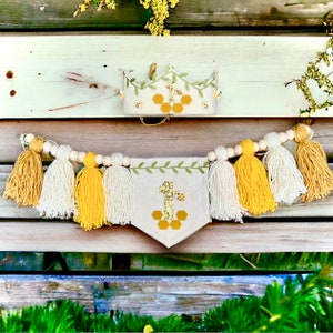 Puede incluir: Una corona y una pancarta de fieltro con tem&aacute;tica de abejas para un primer cumplea&ntilde;os con un dise&ntilde;o de panal, borlas amarillas y blancas y una guirnalda de cuentas de madera.