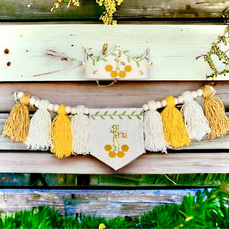 Puede incluir: Una corona y una pancarta de fieltro con tem&aacute;tica de abejas para un primer cumplea&ntilde;os con un dise&ntilde;o de panal, borlas amarillas y blancas y una guirnalda de cuentas de madera.