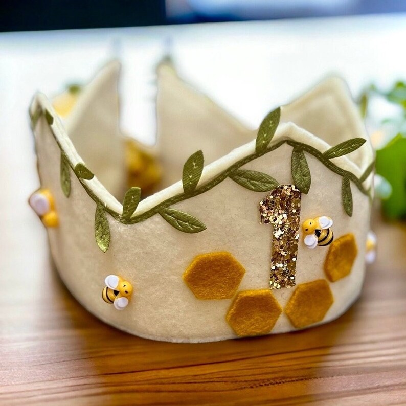 Puede incluir: Una corona de fieltro blanco con una guirnalda de hojas verdes, un n&uacute;mero uno de purpurina dorada y dise&ntilde;os de panal de abeja de fieltro amarillo. Tambi&eacute;n hay tres peque&ntilde;as abejas de fieltro con alas blancas.