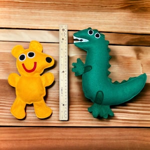 May include: Two felt stuffed toys, a yellow bear with two large eyes and a green dinosaur with a long tail. The toys are on a wooden surface.