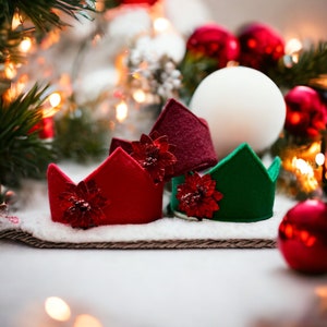 May include: Three miniature Christmas crowns in red, burgundy, and green, each adorned with a red poinsettia. The crowns sit on a white, snowy surface, with Christmas ornaments and lights in the background. Festive holiday decor.
