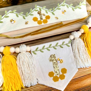 Puede incluir: Una corona de fieltro blanco con hojas verdes y abejas amarillas, una cinta dorada y un panal de abejas de fieltro con un n&uacute;mero uno de purpurina dorada. Un bander&iacute;n blanco con hojas verdes, un n&uacute;mero uno de purpurina dorada, una abeja amarilla y un dise&ntilde;o de panal. El bander&iacute;n est&aacute; unido a una cuerda con cuentas de madera y borlas amarillas y blancas.