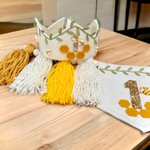 Puede incluir: Una corona de fieltro con hojas verdes y un dise&ntilde;o de panal, decorada con un n&uacute;mero uno de fieltro y peque&ntilde;as abejas. La corona va acompa&ntilde;ada de una pancarta de fieltro con un gran n&uacute;mero uno y un dise&ntilde;o de panal, y un conjunto de borlas de hilo amarillo y blanco.