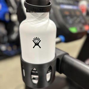 Puede incluir: Una botella de agua Hydro Flask blanca con un logotipo negro está asegurada en un soporte negro sujeto a una cinta de correr. La botella tiene una tapa negra con el texto "Hydro Flask". El soporte está diseñado para la botella.