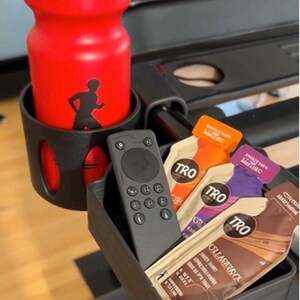 May include: Red water bottle with "PROHYRATE" text and a runner silhouette, placed in a black holder. A black remote and several packets of TRO energy gels are in a tray. The items are on a treadmill.