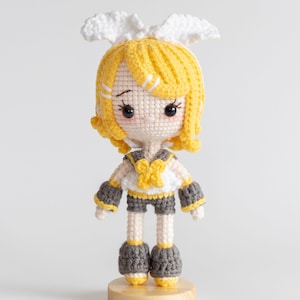 Może przedstawiać: Ręcznie robiona szydełkowa lalka z żółtymi włosami i pasującym strojem. Lalka ma biało-żółty top, szare szorty i szare buty. Lalka stoi na drewnianej podstawie i ma figlarny wyraz twarzy.