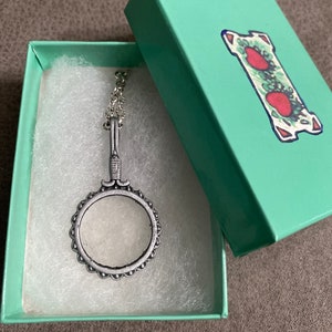 Puede incluir: Un collar de plata con un colgante de lupa en una caja de regalo verde claro con un diseño de fresa.