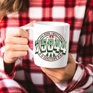 以下が含まれることがあります： 緑と赤のデザインが施された白いセラミック製のマグカップ。マグカップには「Home Sweet 75844 Hometown」の文字が書かれています。