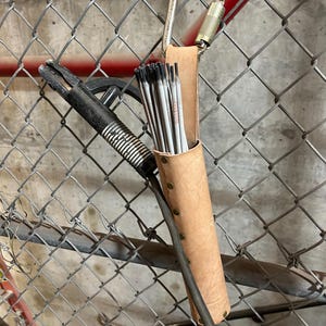May include: A brown leather welding rod holder with a carabiner clip. The holder is filled with welding rods and is hanging on a chain link fence.