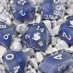 Puede incluir: Un conjunto de siete dados poliédricos con un diseño de galaxia azul oscuro y púrpura. Los dados tienen números blancos y están colocados sobre un fondo de piedras blancas. Algunos dados presentan un icono de tetera blanca.
