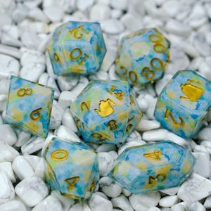 May include: Set of seven polyhedral dice with a blue, yellow, and green marbled design. The dice have gold-colored numbers and a gold teapot icon on one face. The dice are displayed on a bed of white stones.