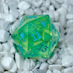 K&ouml;nnte beinhalten: Ein durchscheinender gr&uuml;n-gelber polyedrischer W&uuml;rfel mit blauen Zahlen. Der W&uuml;rfel liegt vor einem Hintergrund aus wei&szlig;en Steinen. Der W&uuml;rfel ist ein Spielzubeh&ouml;r, wahrscheinlich f&uuml;r Tabletop-Rollenspiele.