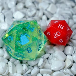 K&ouml;nnte beinhalten: Zwei polyedrische W&uuml;rfel, einer gr&uuml;n und einer rot, liegen auf wei&szlig;en Steinen. Der gr&uuml;ne W&uuml;rfel hat blaue Zahlen und ein blaues Design, w&auml;hrend der rote W&uuml;rfel wei&szlig;e Zahlen hat. Diese W&uuml;rfel werden f&uuml;r Tabletop-Spiele verwendet.