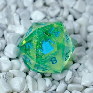 K&ouml;nnte beinhalten: Ein durchscheinender gr&uuml;n-gelber polyedrischer W&uuml;rfel mit blauen Zahlen und einem blauen Teekannen-Symbol. Der W&uuml;rfel liegt auf einem Bett aus wei&szlig;en, glatten Steinen. Es ist ein Spielzubeh&ouml;r.