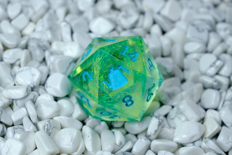 K&ouml;nnte beinhalten: Ein durchscheinender gr&uuml;n-gelber polyedrischer W&uuml;rfel mit blauen Zahlen und einem blauen Teekannen-Symbol. Der W&uuml;rfel liegt auf einem Bett aus wei&szlig;en, glatten Steinen. Es ist ein Spielzubeh&ouml;r.