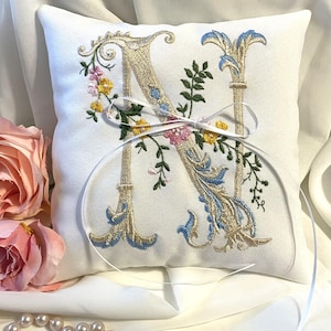 May include: White satin ring pillow with a floral embroidered design featuring the letter "N" in a script font. The pillow is tied with a white satin ribbon.