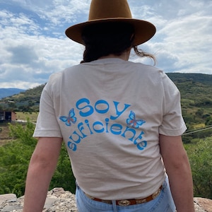 May include: A person wearing a light brown fedora and a beige t-shirt with the text "Soy Suficiente" in blue with two blue butterflies on either side of the text. The person is wearing blue jeans and a brown belt with a gold buckle.