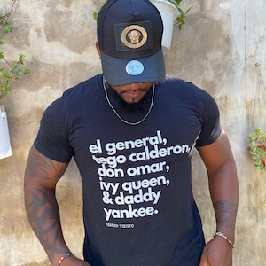 May include: Black t-shirt with white text that reads "el general, tego calderon, don omar, ivy queen, & daddy yankee." The text is centered on the shirt. The shirt also has the text "PERREO VIEJITO" in smaller white text at the bottom. The person wearing the shirt is also wearing a black mesh baseball cap with a gold Versace logo.