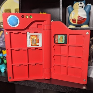 May include: A red Nintendo 3DS game card case with a built-in game card holder. The case has a blue button and a clear plastic window to display the game card. The case is open and shows a Nintendo 3DS game card with the text "New Super Mario Bros. 2" on it.