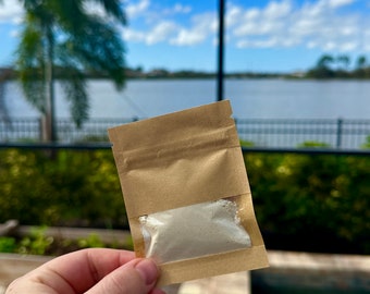 May include: A small brown paper pouch with a clear plastic window, containing a white powder. The pouch is being held in a hand, with a pool and palm trees in the background.