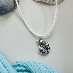 May include: A silver-colored seashell pendant necklace with a white cord. The pendant is a detailed spiral design. The necklace is displayed on a white surface with seashells and a light blue scarf.
