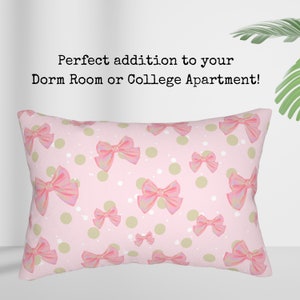 Op de afbeelding: Roze rechthoekig kussen met een patroon van roze strikken, groene stippen en witte spikkels. De tekst op het kussen luidt "Perfect addition to your Dorm Room or College Apartment!"
