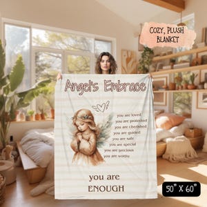 May include: A white plush blanket with the words "Angel's Embrace" and a quote about being loved, protected, cherished, guided, safe, special, precious, and worthy. The blanket also features a watercolor illustration of an angel with wings and a heart. The blanket is 50 inches by 60 inches.