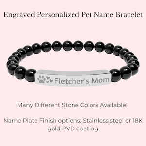 Pode incluir: Pulseira de contas pretas com uma placa de identificação prateada. A placa de identificação está gravada com "Fletcher's Mom" e pegadas de patas. O texto "Engraved Personalized Pet Name Bracelet" está no topo. O texto "Many Different Stone Colors Available!" está abaixo.