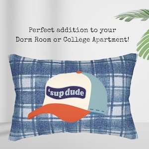 May include: Rectangular cushion with a blue plaid design. The cushion displays a baseball cap graphic with the text "'sup dude". The cap has a white front, blue bill, and orange brim. The text "Perfect addition to your Dorm Room or College Apartment!" is above the cushion.