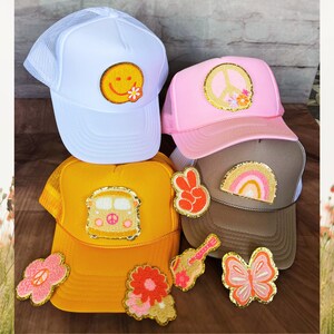 Puede incluir: Cuatro gorras de camionero con varios parches, incluyendo una cara sonriente, un signo de paz, un arcoíris y una camioneta vintage. Las gorras son blancas, rosas, marrones y amarillas. También hay parches adicionales, incluyendo una guitarra, una mariposa y una flor.