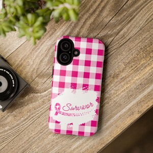 Op de afbeelding: Roze en witte geruite telefoonhoes met een wit splatterontwerp en een roze lint met het woord "Survivor" in witte tekst.
